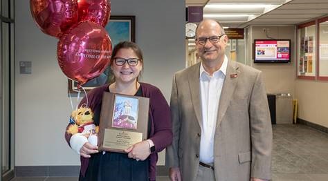 Kate Peffley smiling and holding her Employee of the Month placard, balloons, and stuffed golden bear next to President Hawkinson, who is smiling 