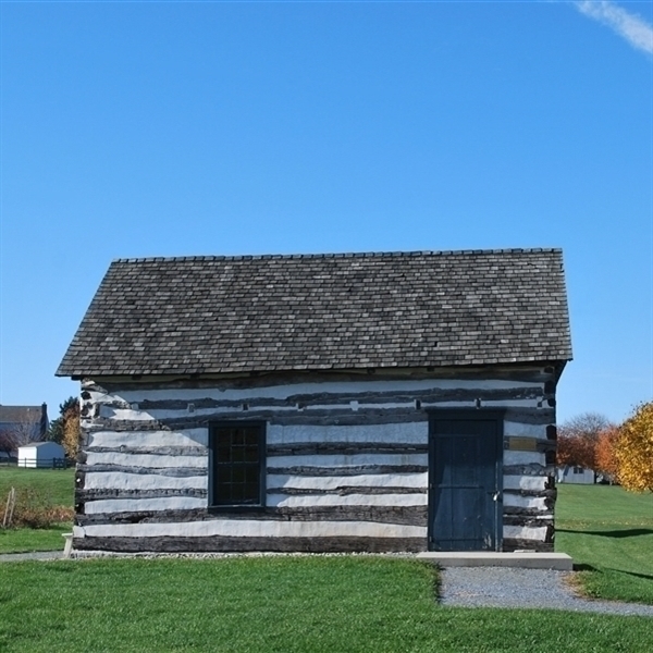 Image of a log cabin with a shingle roof against a blue sky. There is white morter between the hand hewn logs
