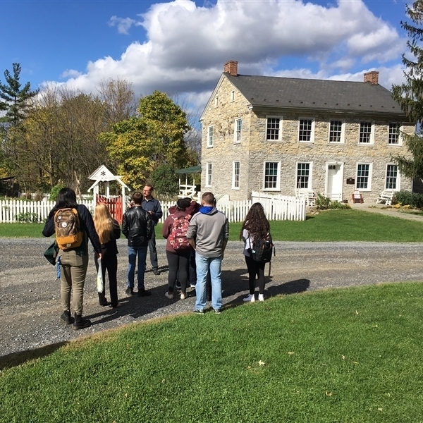 A tour group of students standing in the gravel driveway in front of the house, listening to the tour guide.