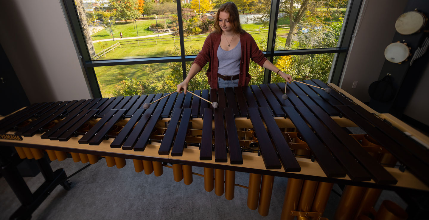 image of a white female student standing infront of a window showcasing the landscape of campus also playing a marimba