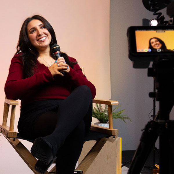 Female communication studies major sitting in a directors chair, holding a microphone. In the foreground is a camera's viewfinder, that shows another angle of the student.
