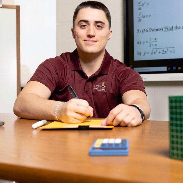 Male math education major holding a pen above a pad.  In the foreground is a blue calculator, green box with supplies and a laptop computer.  In the backgrround are math formulas and questions.