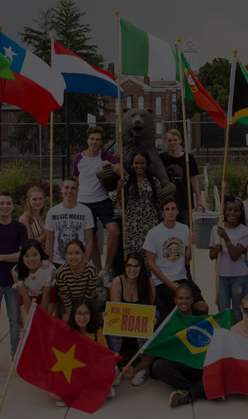 International students at Kutztown University with the flags of their countries arranged around the Golden Bear statue.