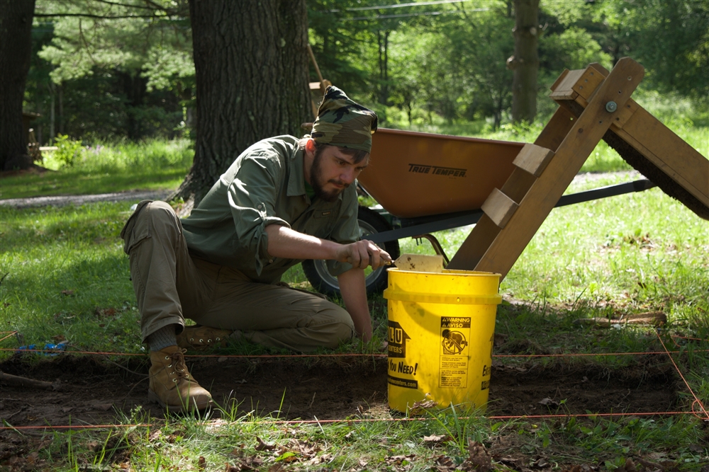 Student collecting soil from a taped-off area and depositing it in a bright yellow bucket
