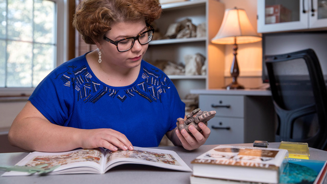 Female student studying an artifact while looking at a reference guide.