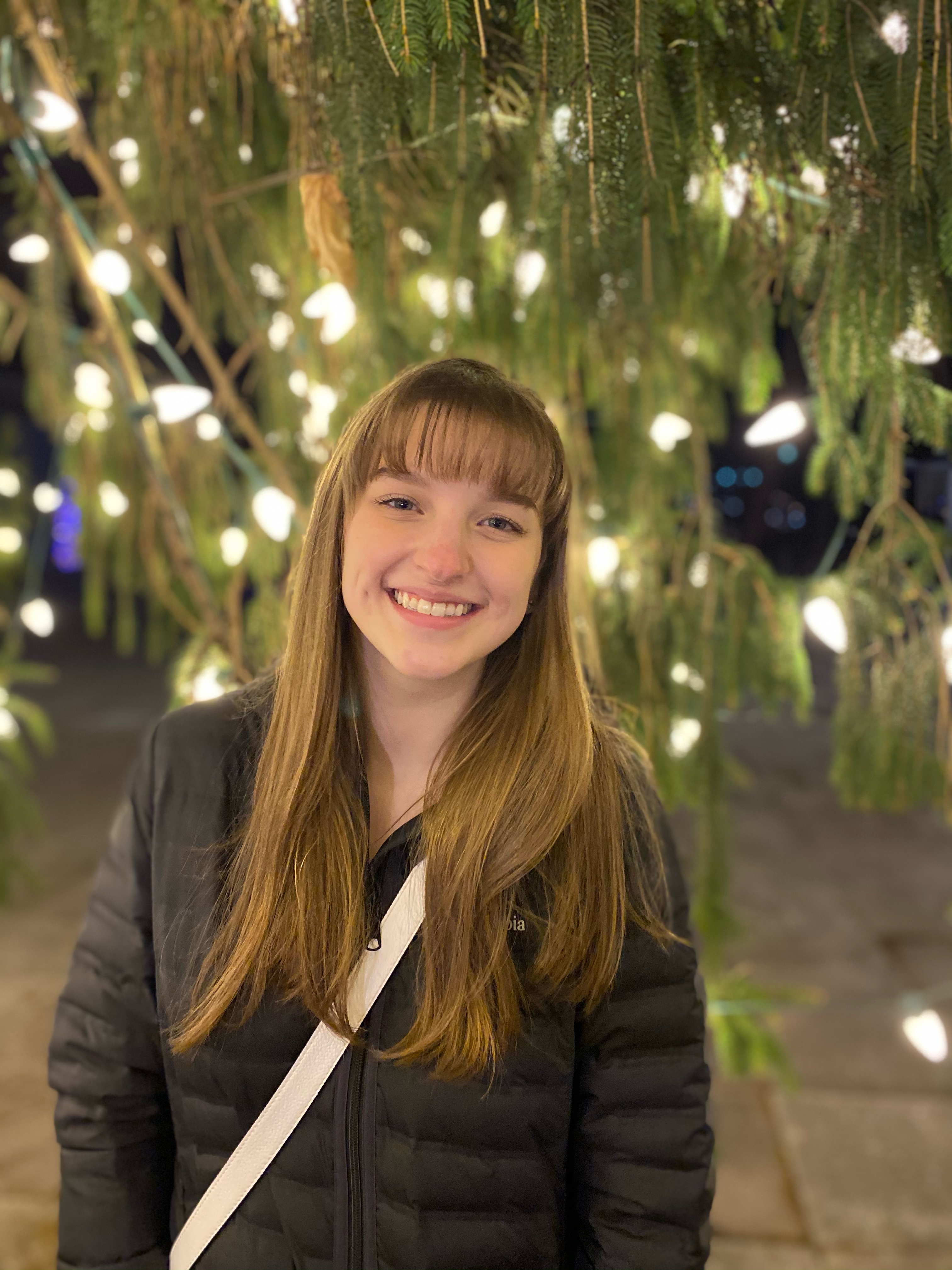 Joclyn Rodick headshot, with her smiling in front of a tree decorated with Christmas lights 