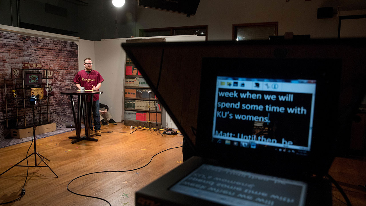 Closeup of a teleprompter with a male student reading into a microphone in the background 