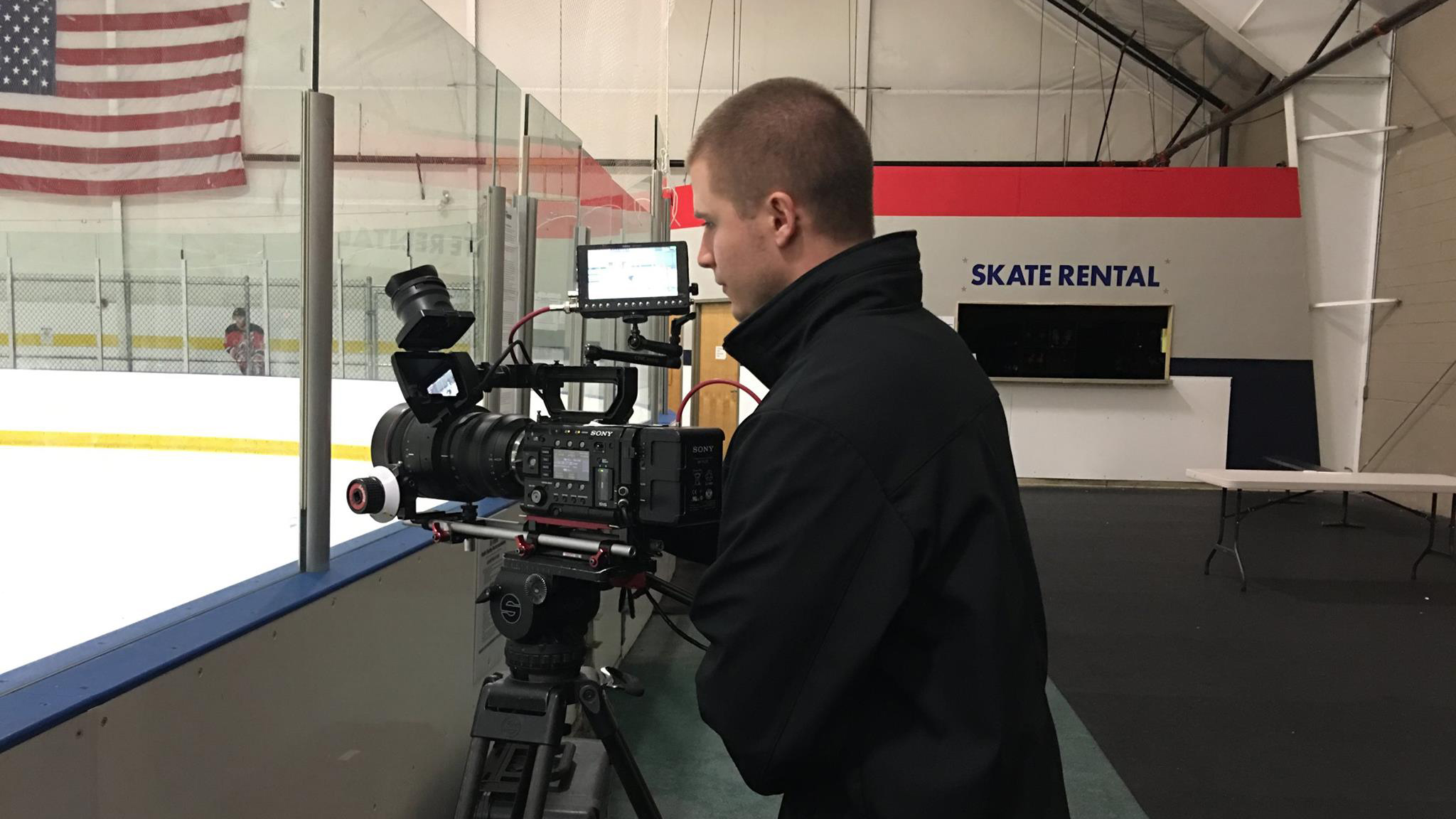 Male student shooting with large camera at an ice hockey rink with a sign that says "skate rental" in the background 