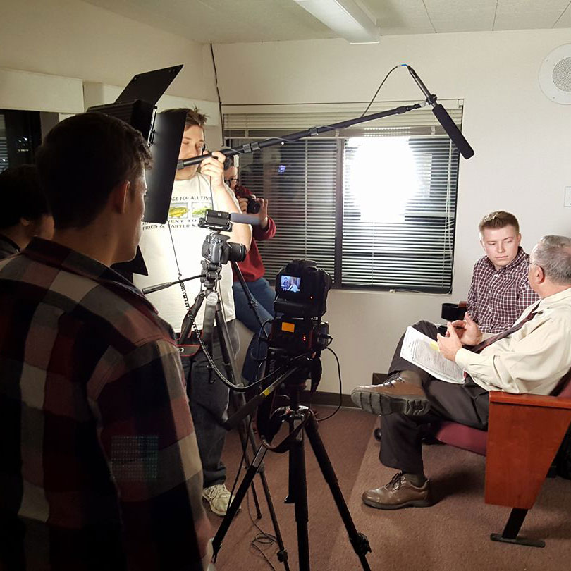 KU-TV crew setting up filming equipment, including cameras and lighting, in front of interviewer and interviewee, who sit in a pair of red armchairs across from the equipment 