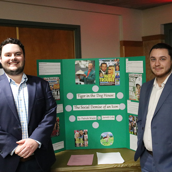 Two students standing on either side in front of a poster presentation titled "tiger in the doghouse: the social demise of an icon." 