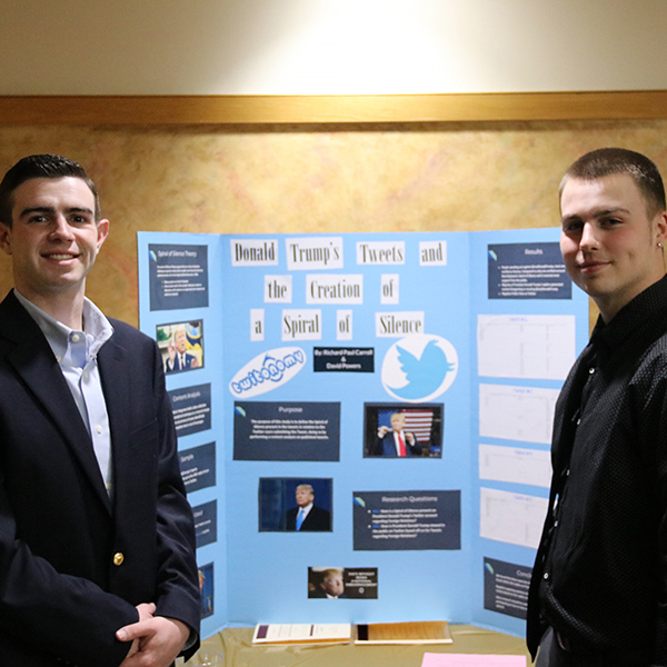 Two students standing on either side in front of a poster presentation titled "Donald Trump's tweets and the creation of a spiral of silence." 