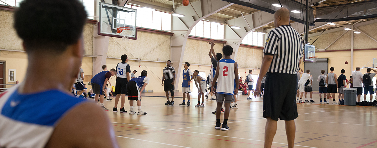 Wide shot of youth basketball practice game, with two referees and two small teams watching a young man shoot at the basket