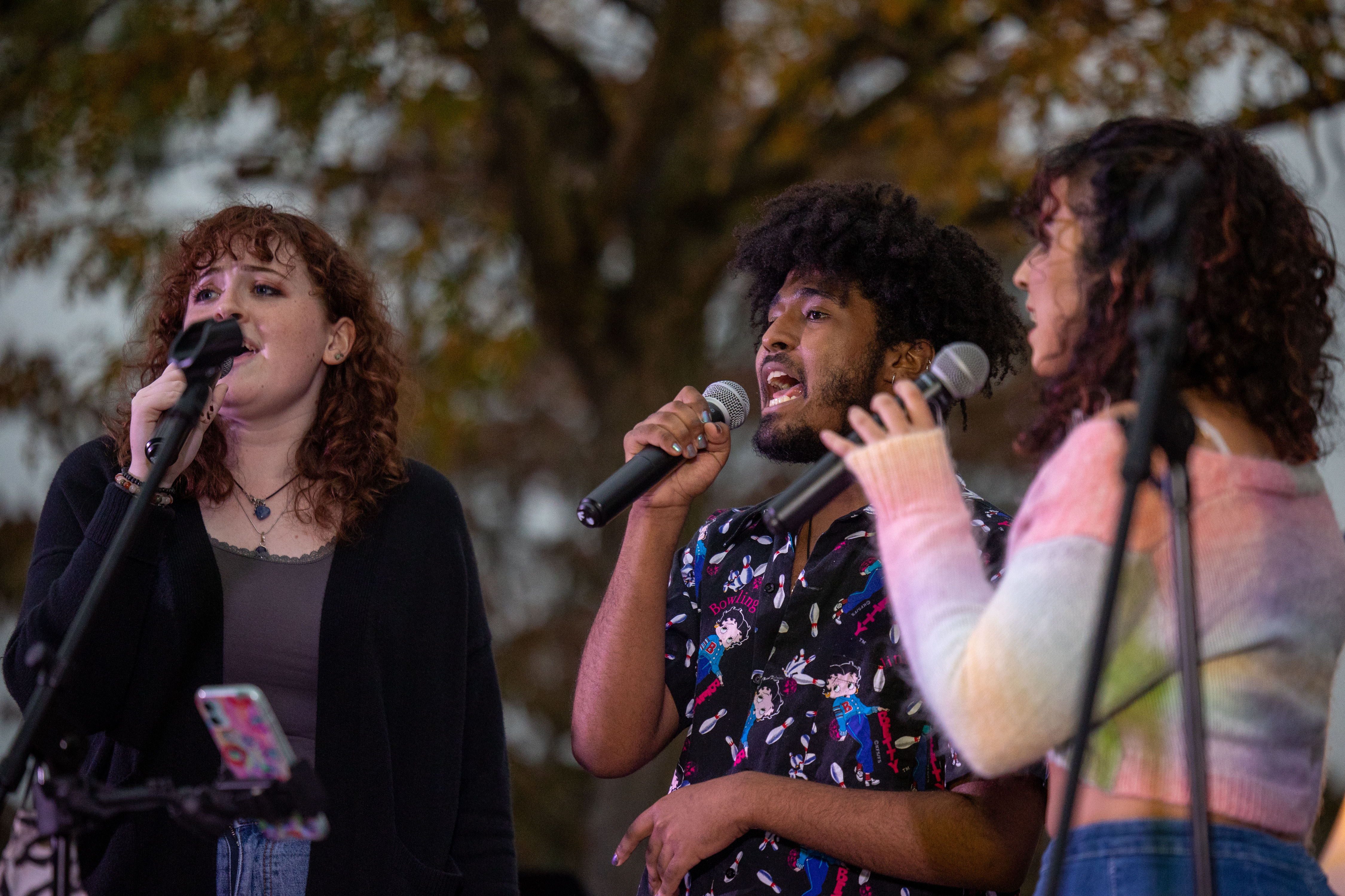 3 students holding microphones and singing outside in fall