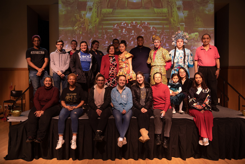 group of 20 people sitting on stage in front of projector screen indoors