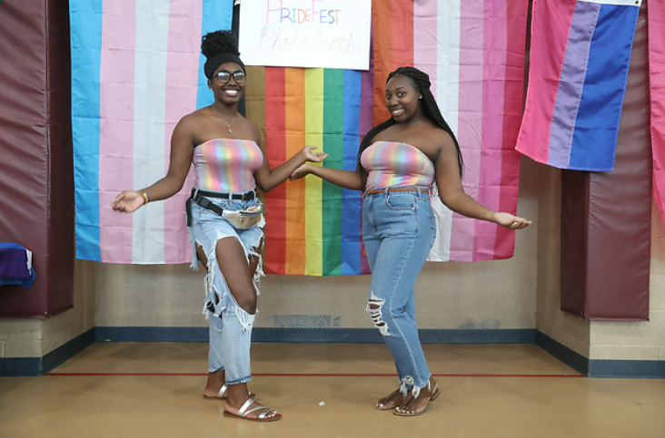 2 female students standing in front of pride flag indoors