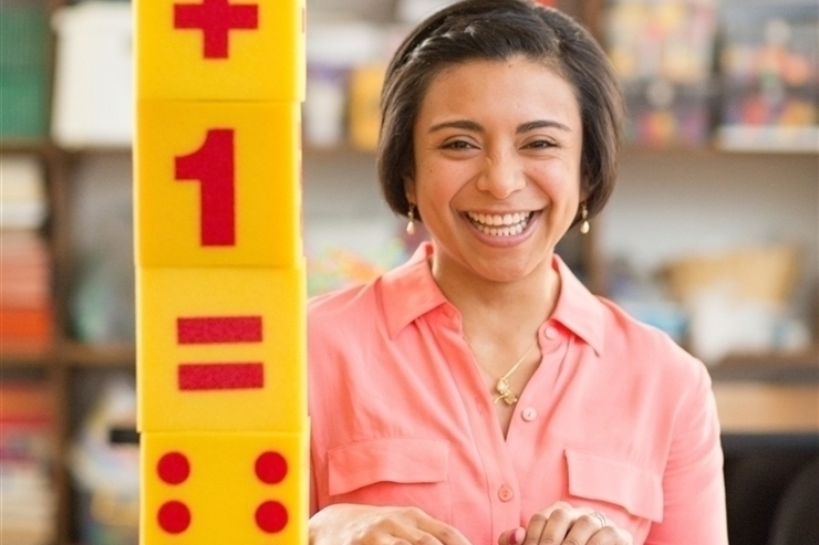 Female student smiling next to a stack of large yellow blocks imprinted with mathematic symbols