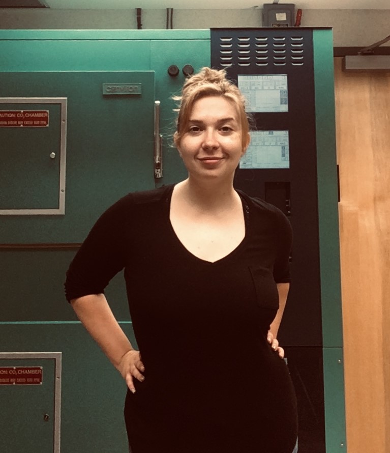 Emily Bohn wearing black v-neck shirt standing in front of large green research equipment