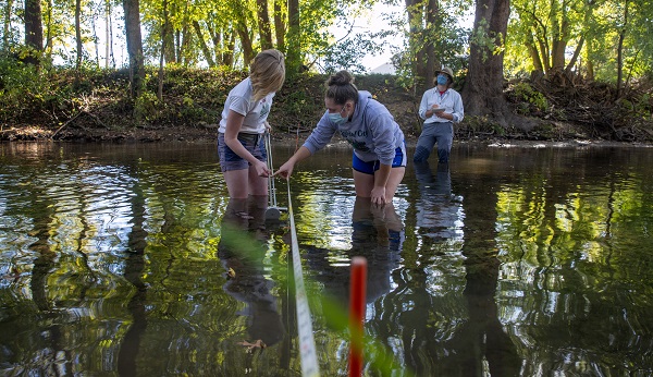 students wearing waders taking discharge measurements in a stream with a tape measure and metal rod