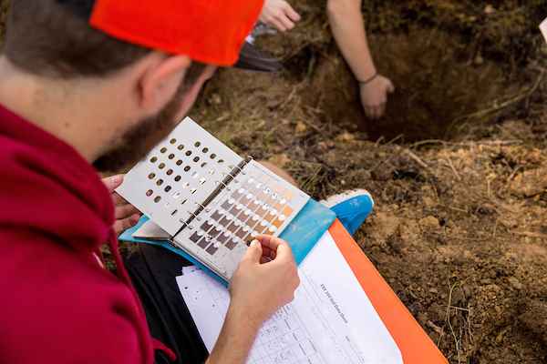 student wearing red sweatshirt and orange cap looking through Monsell soil chart with soil pit in background