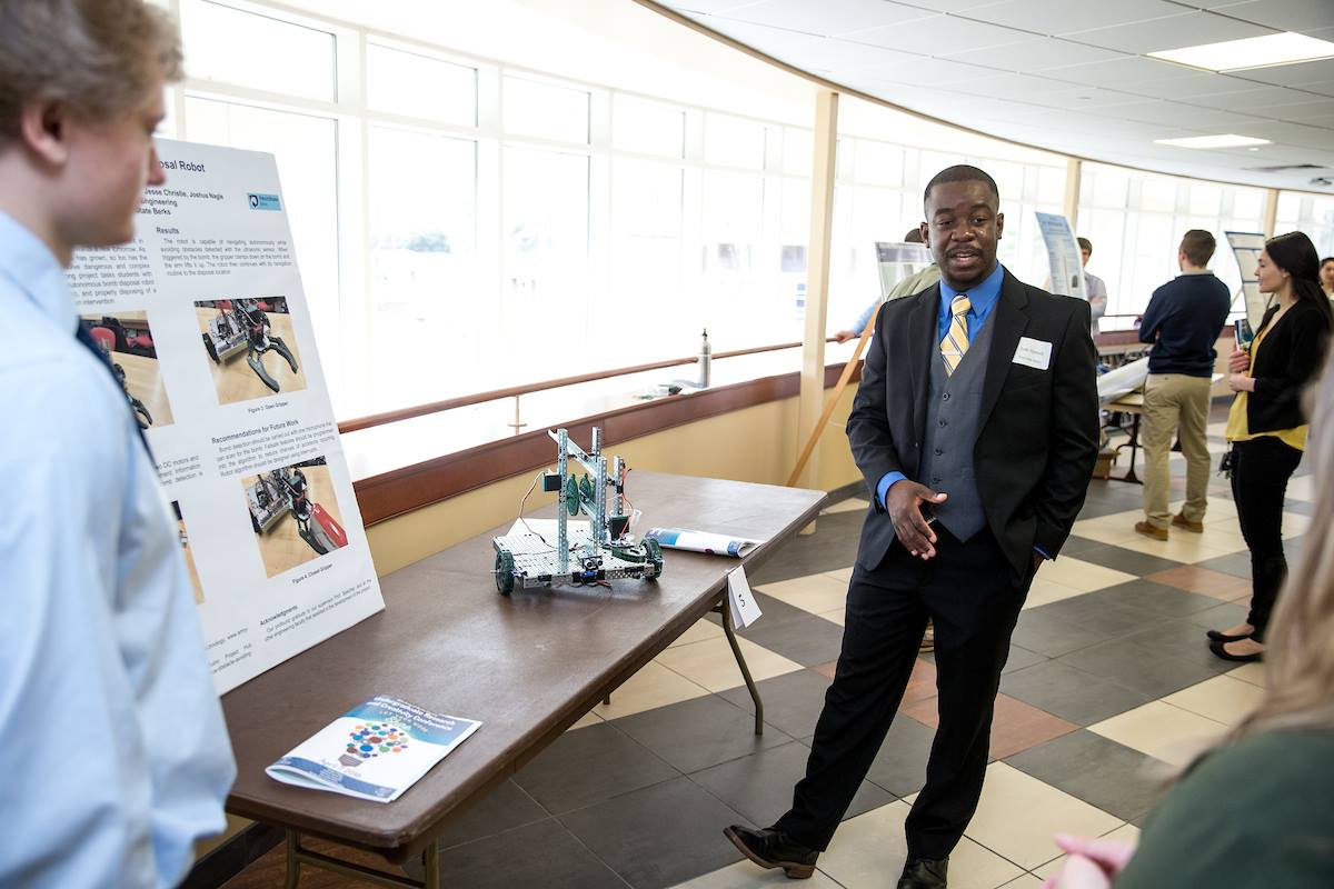 In suit a black suit with yellow and blue striped tie, undergraduate student researcher demonstrates small robot with wheels on a table in front of his detailed poster. 