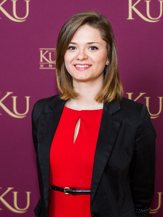 Rebecca Van Horn smiling and standing in front of a KU wallpaper backdrop 