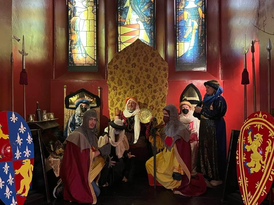 Costumed actors posing in a lavish gold and red throne room, dressed in historical garments, with knights kneeling in front of a king and other royal attendees sitting on thrones beside the king 