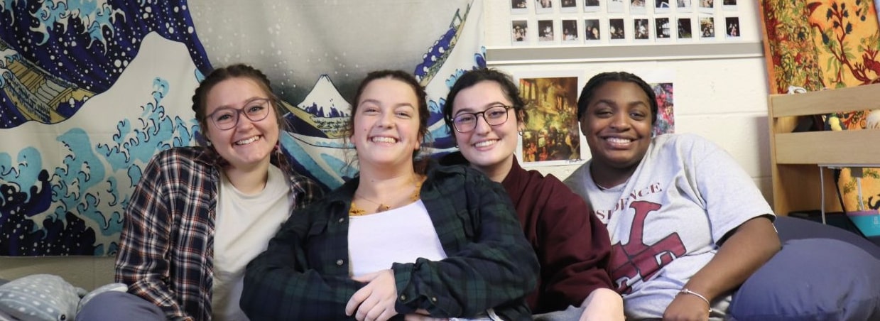 Four residents sitting on residence hall bed smiling for camera