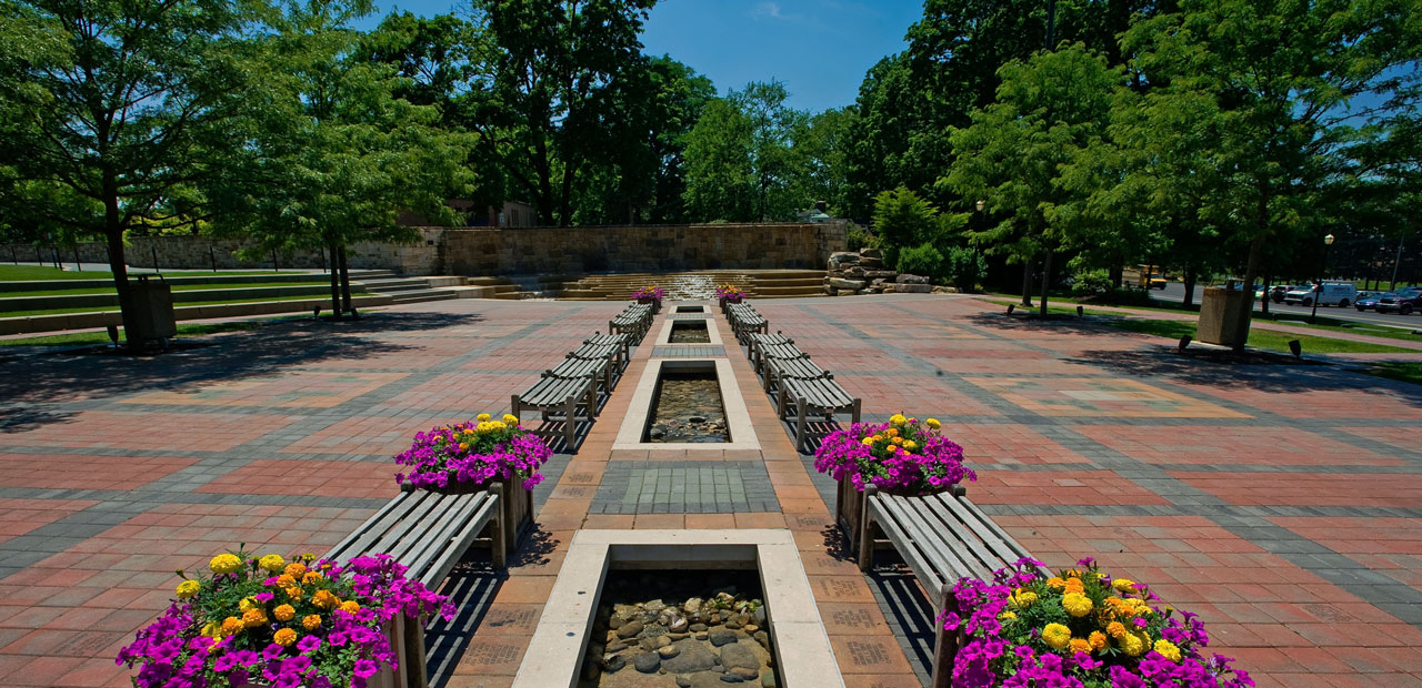 Wide shot of Alumni Plaza in the spring, with flowering plants in between the benches leading up to the main fountain 