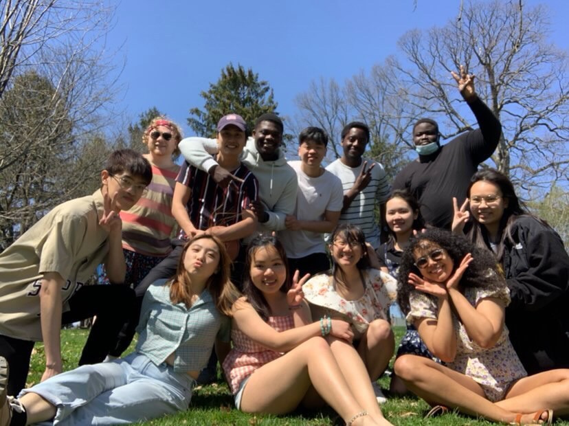 Large group of students smiling and sitting on the grass together, blowing kisses and waving their hands 