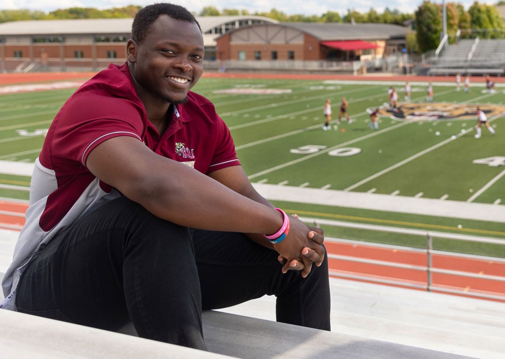 Male student smiling and sitting sideways on bleachers with his hands folded in front of him, with the football field and players practicing in the background 