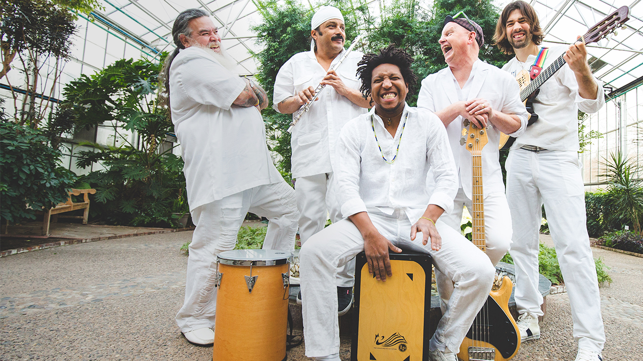 Male band members dressed in white and standing as a group with their instruments, smiling and laughing with each other