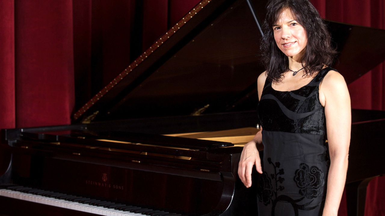 Dr. Maria Asteriadou leaning against a grand piano on stage in an auditorium 