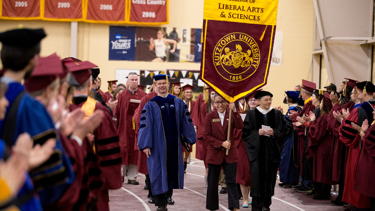 Female student carries the LAS College seal at Graduation Ceremony, with graduating students and LAS faculty in ceremonial robes in a crowd around her