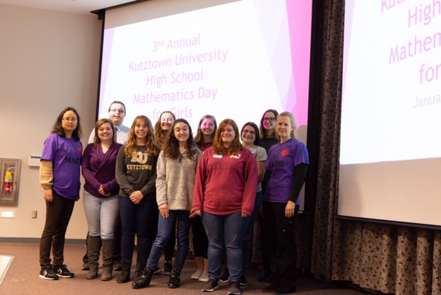 2019 Team smiling on a stage in front of a slide that says "3rd Annual Kutztown University High School Mathematics Day for Girls." 