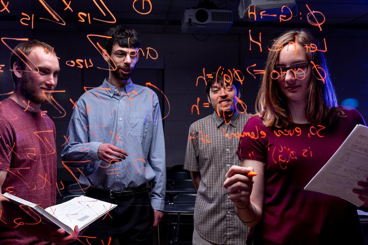 Students and faculty writing and studying mathematics written in orange on a glass panel in the foreground