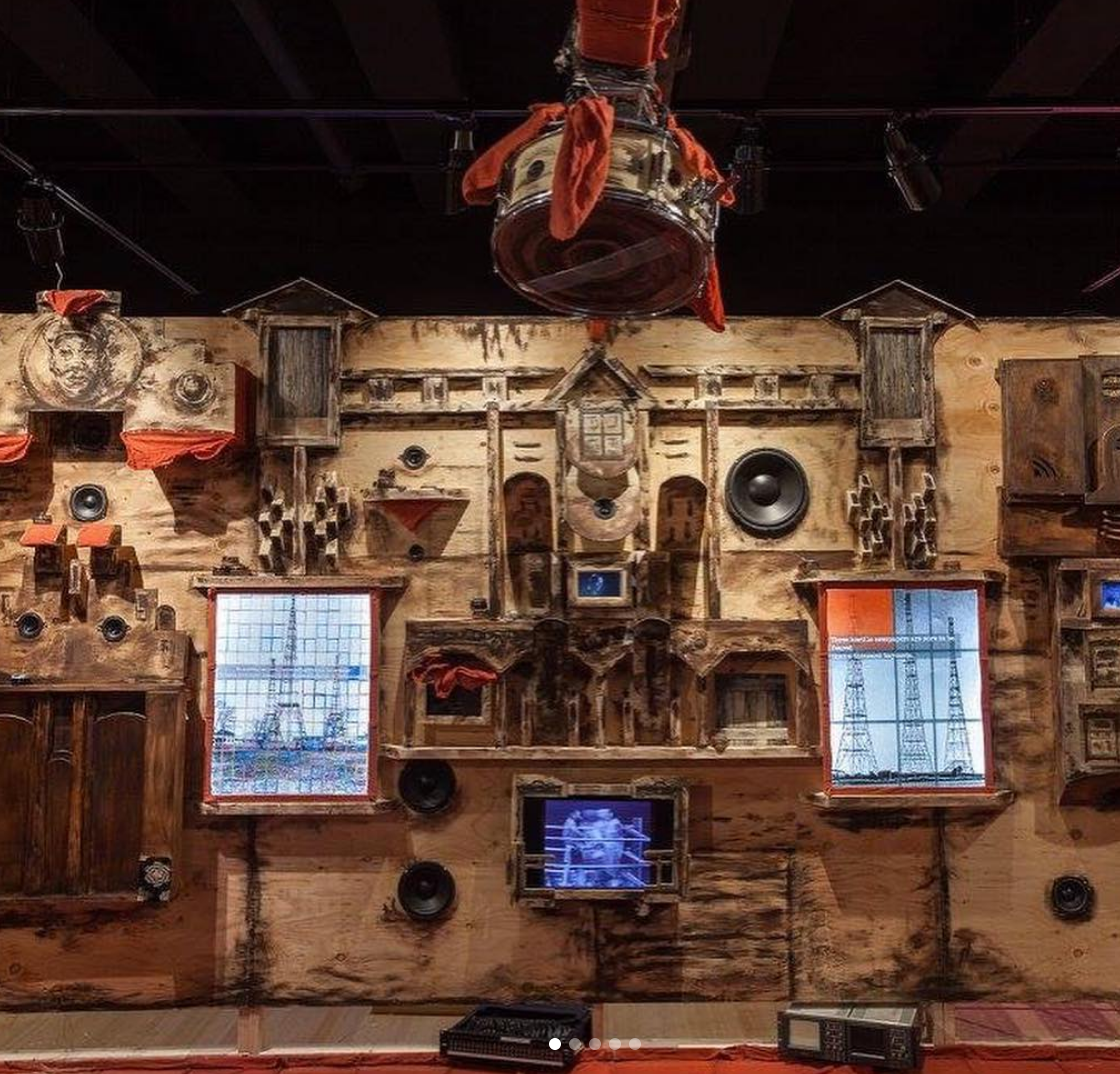 Cultural installation decorated with carved wood pieces and speakers dotted along the wall