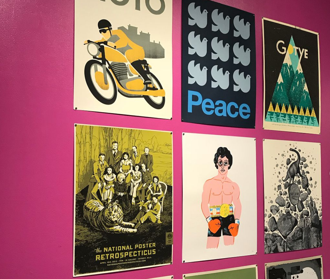 Wall decorated with six posters (from top left to bottom right) of a man on a motorbike, several cartoon doves above the word peace, a Gotye album cover, "the national poster retrospecticus," Rocky Balboa wearing shorts and boxing gloves, and a dinosaur standing on a crumbling stack of rocks  