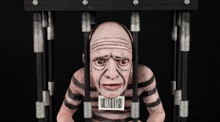 small sculpure of a bald man inside an miniature prison cell, a qr code hangs from his neck and striped are painted on his skin