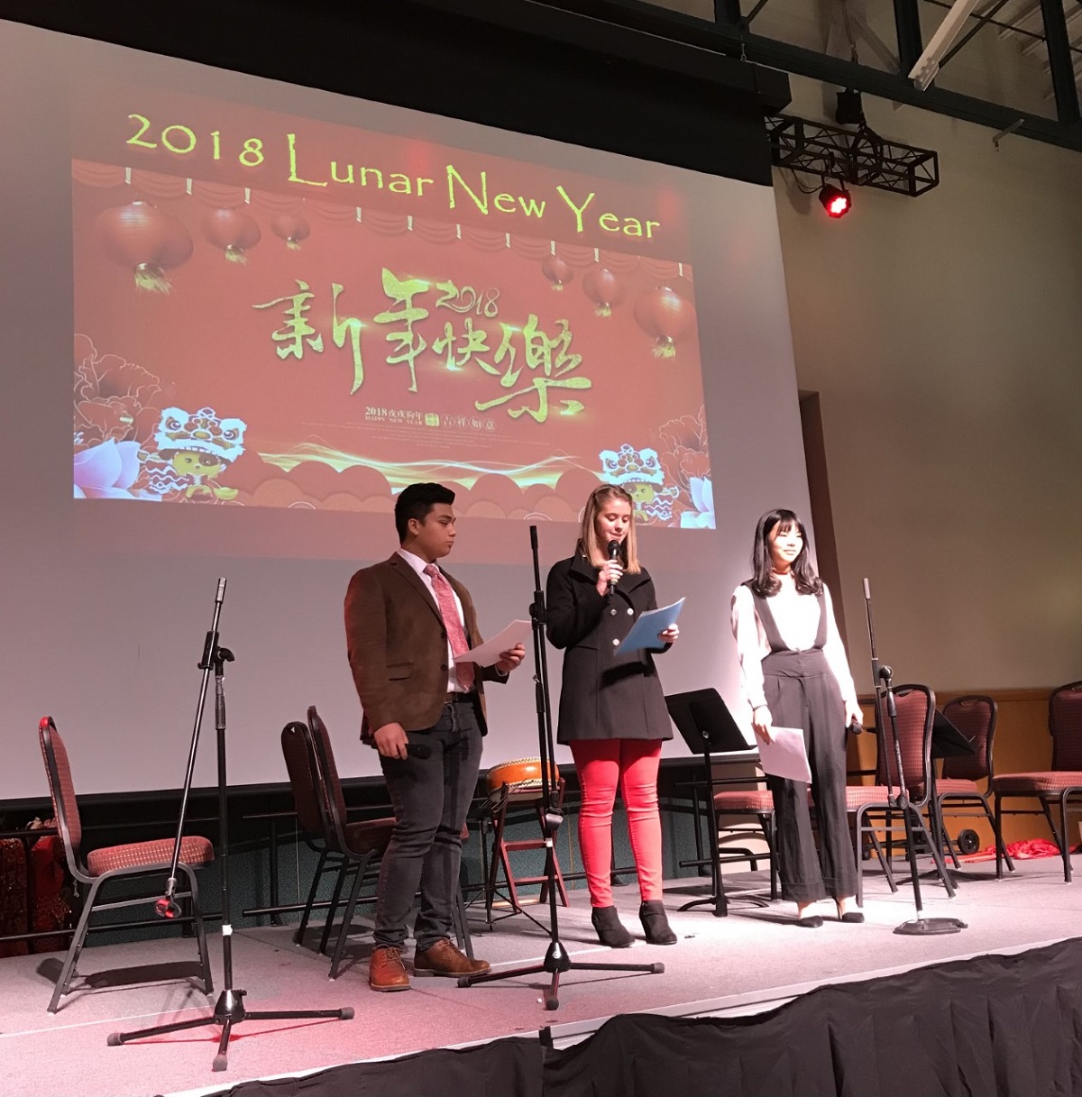 Three students on stage, speaking into a microphone and standing in front of a projection screen with a slide that says "2018 Lunar New Year" in English and Chinese 