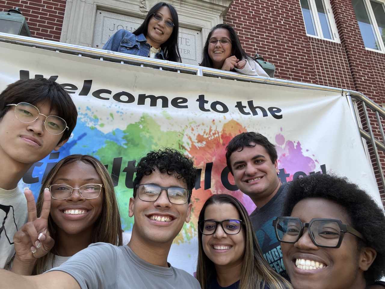 6 students below, 1 student and staff member above MCC welcome banner outside MCC building.