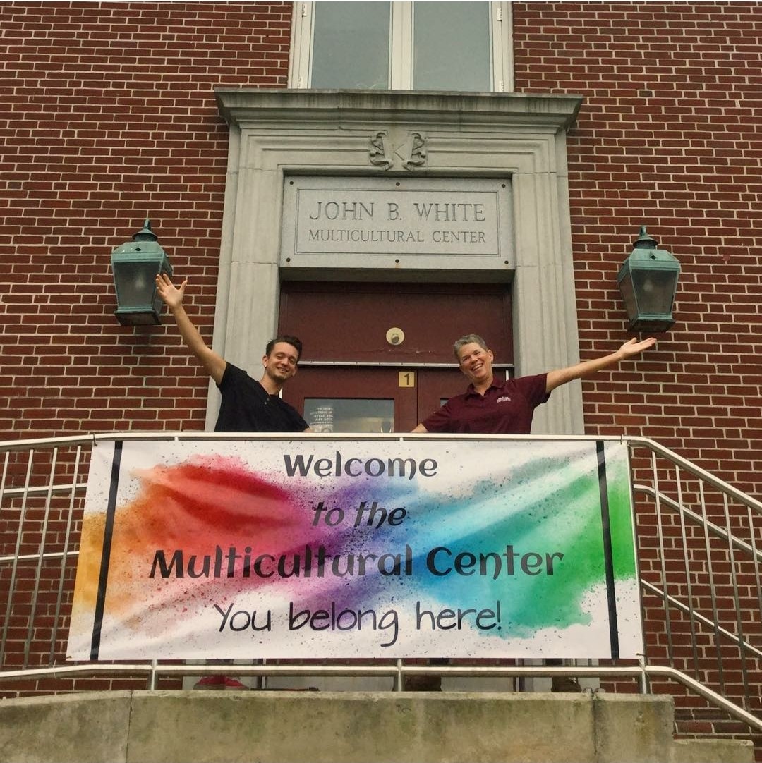 Two people in front of the main door to the Multicultural Center behind a banner that says "Welcome to the Multicultural Center. You Belong Here!" over rainbow watercolor blobs.