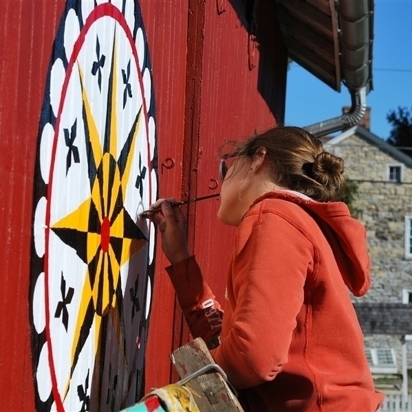 A female identifying student with her head tilted to the right, stands on a ladder painting a yellow, black, white, and red barn star