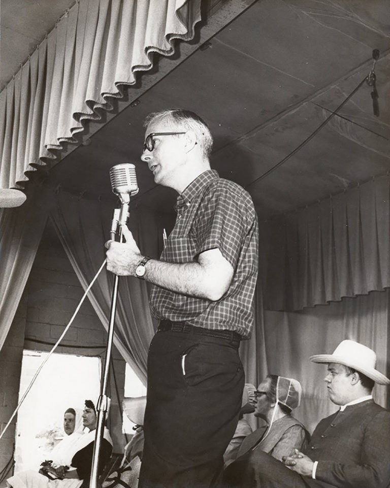 A black and white photo of a white man with white hair stands at a microphone on a stage. He is wearing a checkered shirt and dark pants.