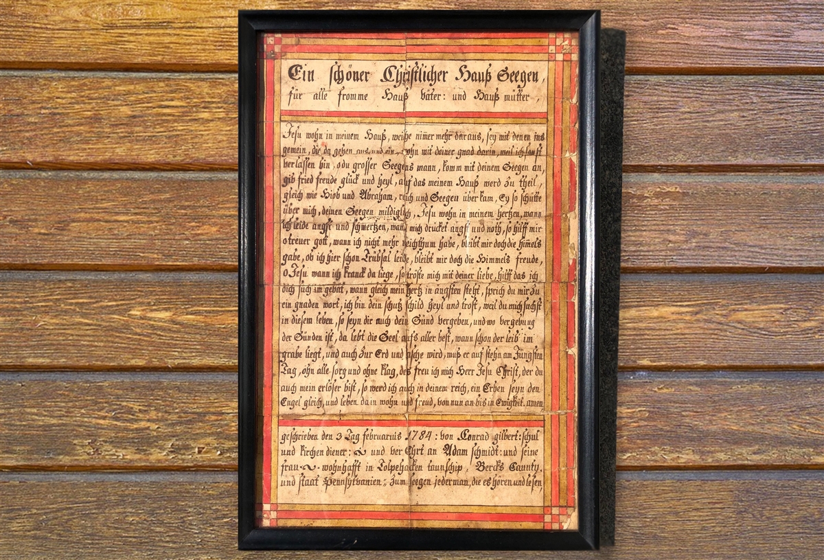 A rectangular document in a black frame, this house blessing is handwritten in dark ink with German script. The border of the document has red and gold detailing.