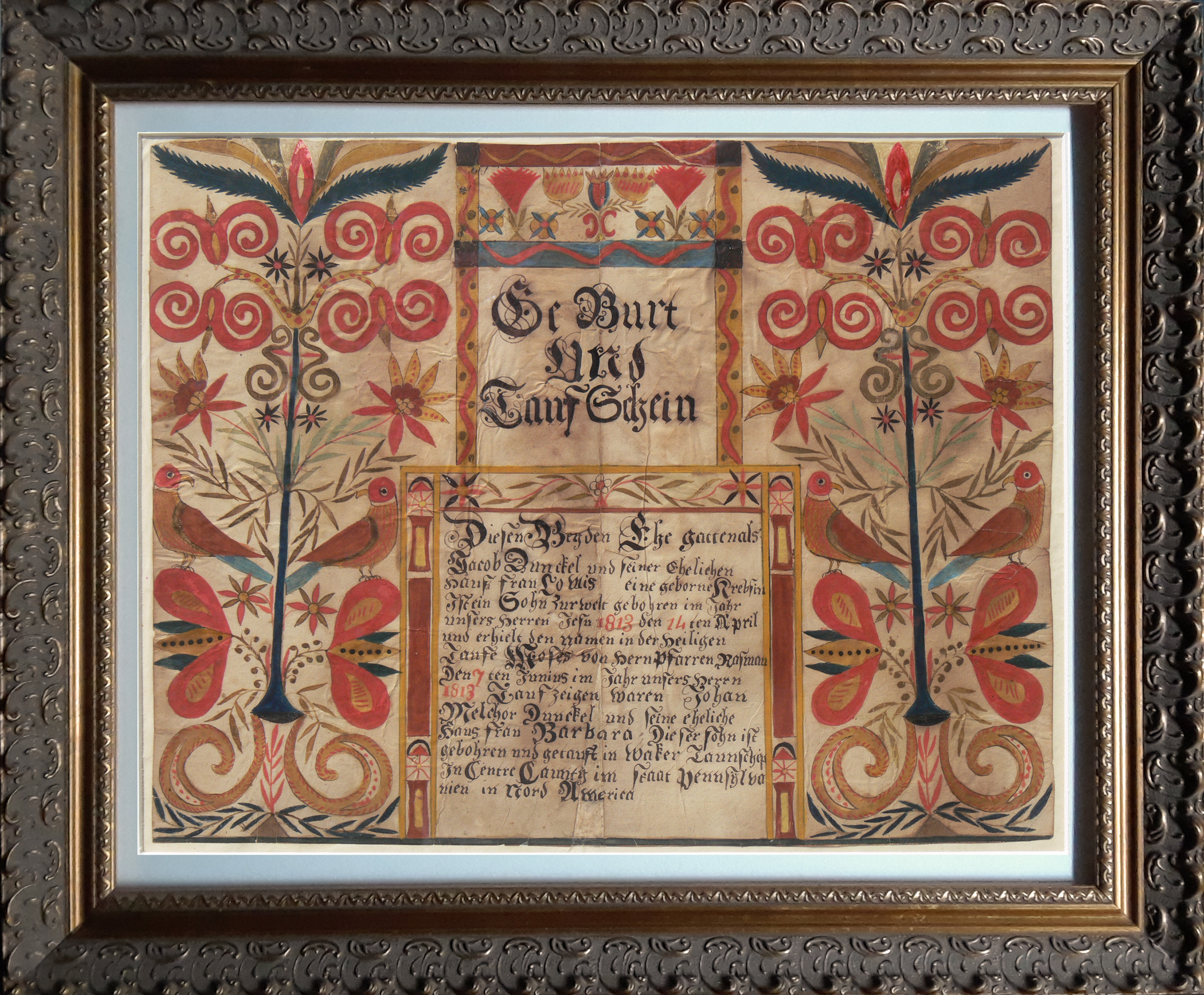 This baptismal certificate is held in an ornate frame. The document has dark ink German script, with important dates written in red ink. There are two sets of birds on either side of the centered text. They are colored in red, blue, and shades of yellow. These birds sit on floral motifs in similar colors, with additional flower and leaf motifs above. The centered text is bordered with boxes decorated columns, zigzags, and flowers.