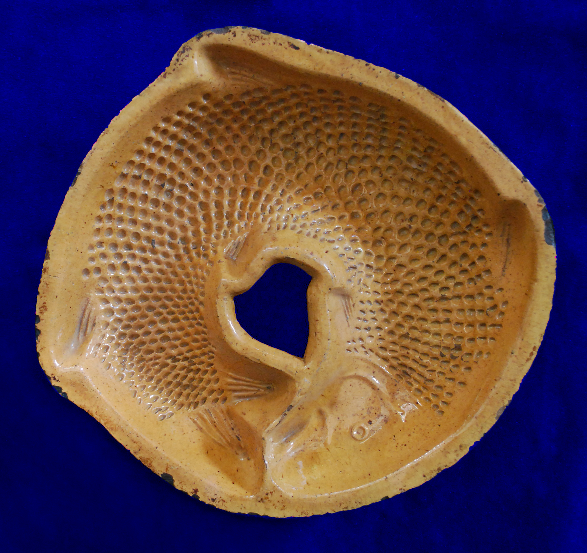 An ocher colored redware dish in the shaped of a fish. The fish’s head meets up with the tail, making the mold a shallow circular shape. There are scales indented into the body, along with fins and facial features. 