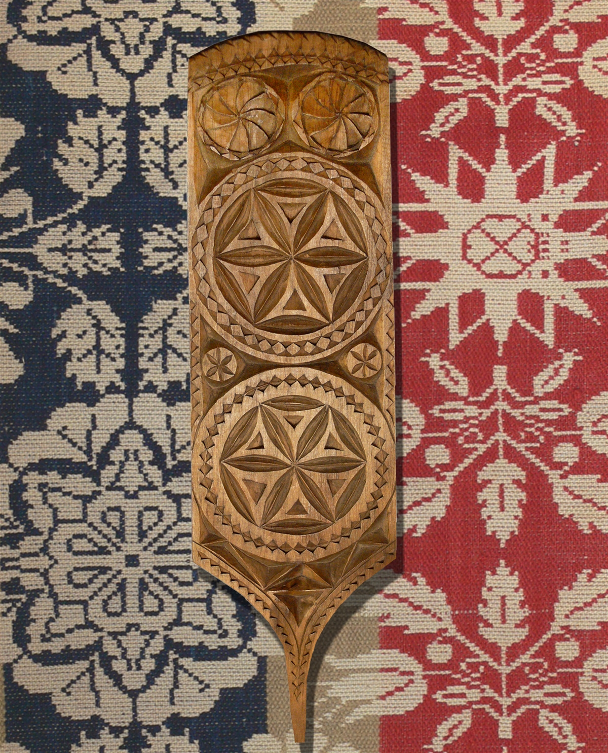 In the foreground of the image is a wooden paddle with a thin handle. The main body has two large 6-pointed chip-carved rosettes. There are two small 6-pointed rosettes between then, and two swirls at the top of the paddle. The border around the paddle is a sawtooth design. In the background is a woven coverlet. There are sections of blue, white, and red, with a repeating pattern of white stars, flowers, and leaves.