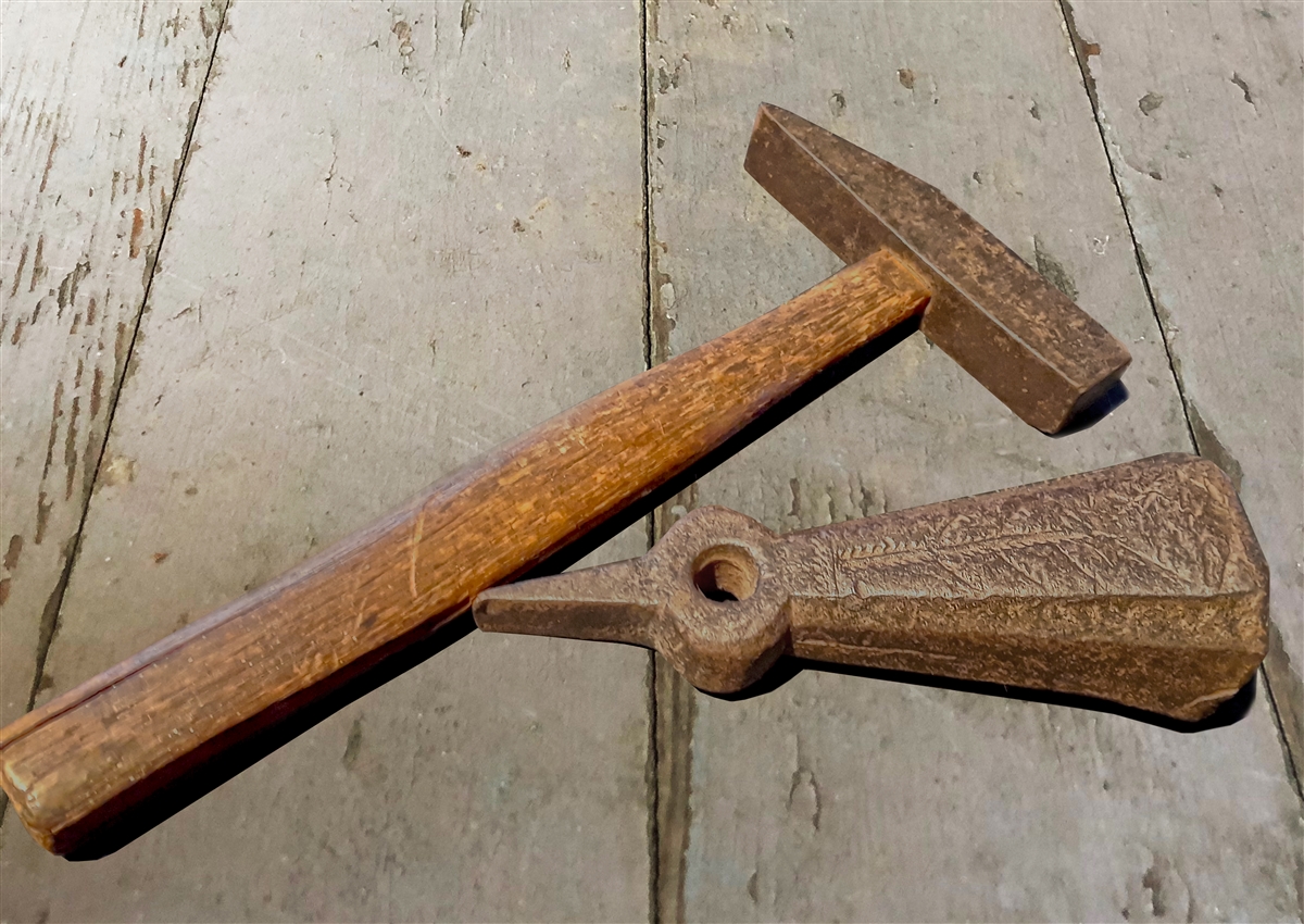 There is a dengelhammer with a small metal head and wooden handle, along with a metal dengelschtock. The dengelschtock has a pointed tip on the one side, and a large faceted end on the other. The larger end in has faint carvings.