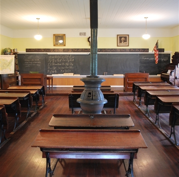 Looking at the slate board at the front of the class from the back of the schoolhouse. Wooden 2-seater desks make three rows facing front.