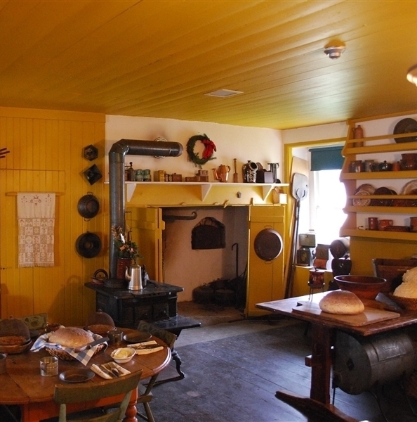 A bright yellow kitchen featuring a hearth, cast iron stove, redware, and other implements on two wooden tables.